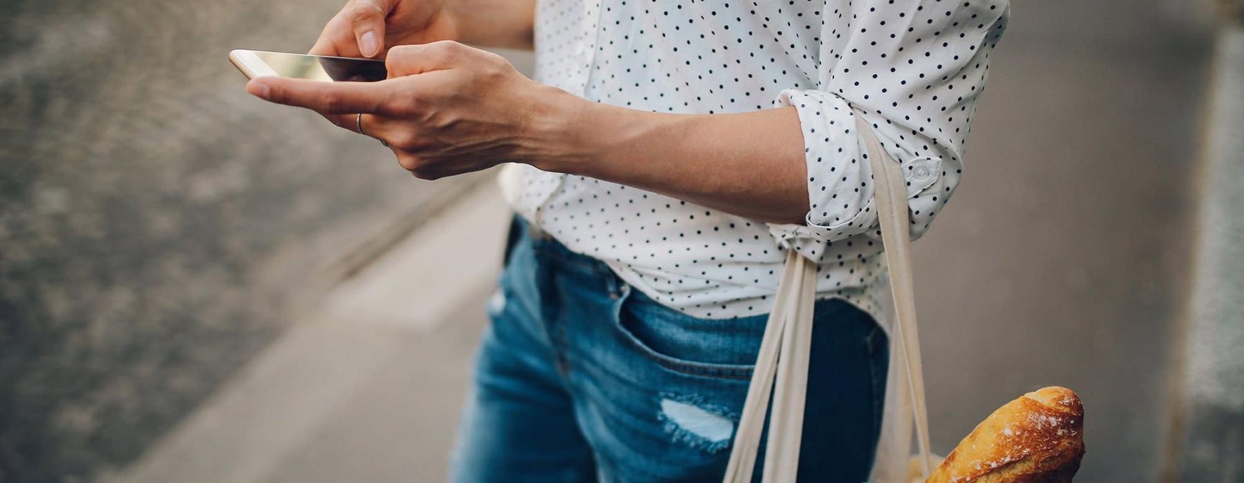 woman walks down the street and texts with a bag of groceries on her arm